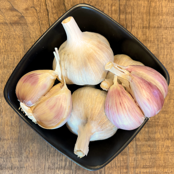 Several whole garlic bulbs with pale ivory skins and pinkish-purple tips arranged in a small black square bowl on a wooden tabletop.